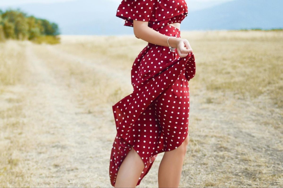 Woman in a red polka dot dress stands on a field, holding her dress's skirt.