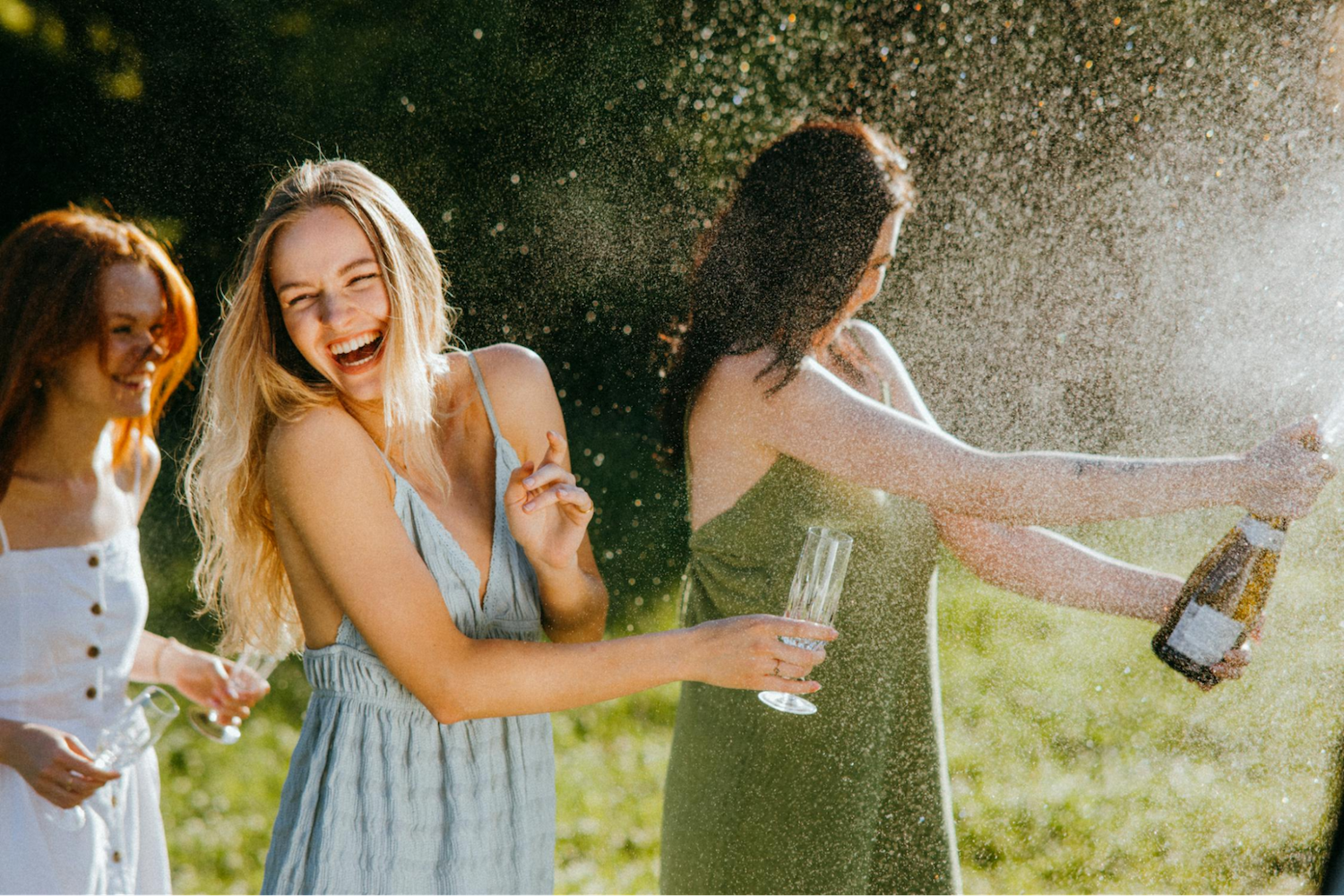 Three women outdoors, laughing as one pops champagne from a bottle.